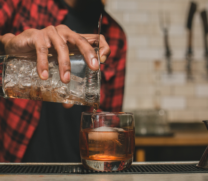 Person pouring amber liquid over ice cubes into a glass on a bar.