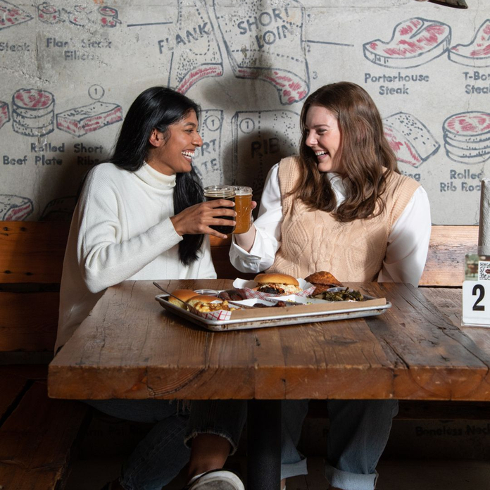 Two women share drinks and food at a rustic wooden table in a restaurant with wall art of meat cuts.