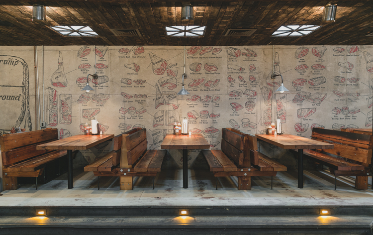 Wooden bench tables and chairs under a wood-paneled ceiling; wall art featuring food-related illustrations.