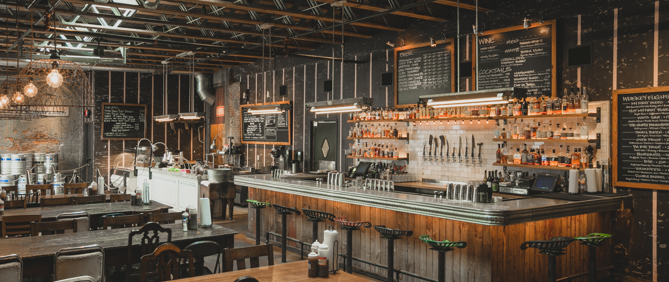 Industrial-style restaurant with wooden tables, exposed metal beams, and a bar with bottles and chalkboards.