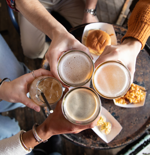 Four hands clinking glasses of beer with food on the table.