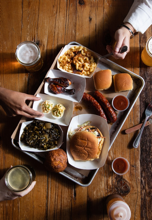 A tray of food featuring mac and cheese, baked beans, sausages, biscuits, grilled chicken, and sides, served with beer glasses.